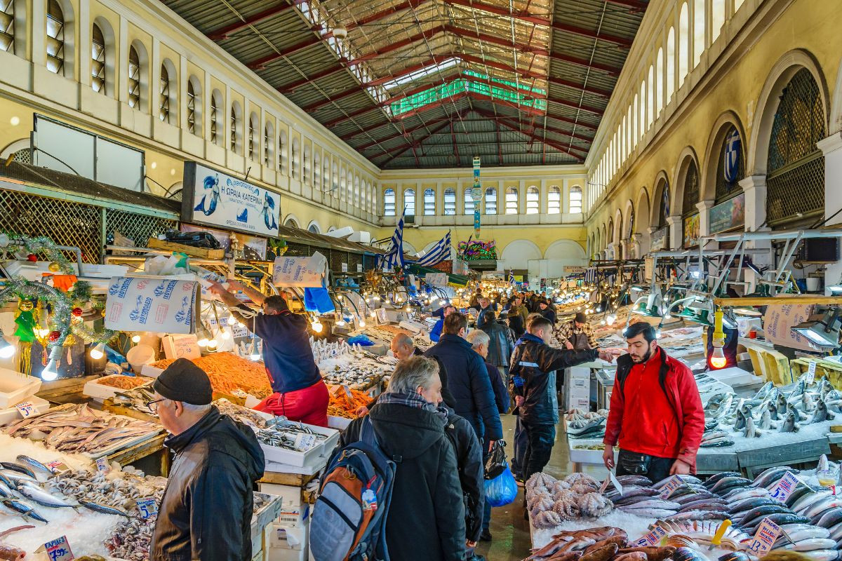 A bustling scene at the Athens fish market, with vendors and customers interacting amidst a variety of fresh seafood displayed on ice, under the market's high, arched ceiling.